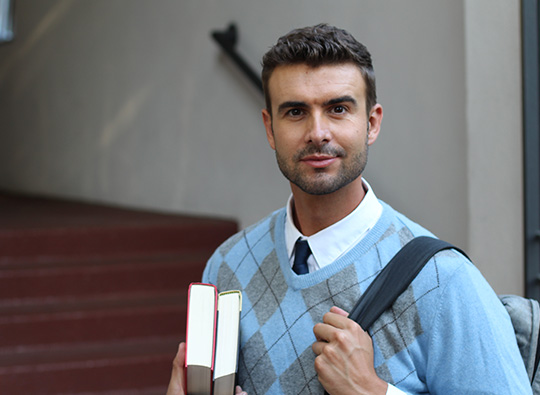 An image of a serious young man in a smart jumper clutching a pile of books.